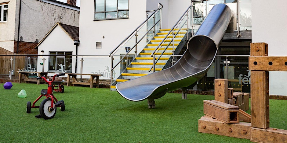 A playground with a shiny metal slide and a red tricycle on green artificial grass, surrounded by wooden play structures and the back of a white building.
