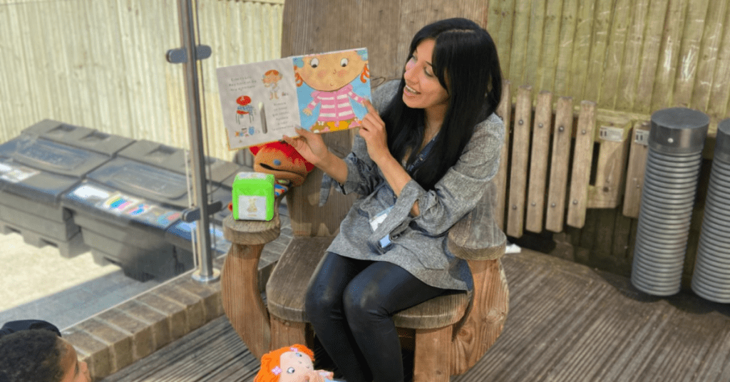 A woman sits outdoors reading a book to children, holding up the book with a colorful cartoon character on the cover, surrounded by toys and listening kids.
