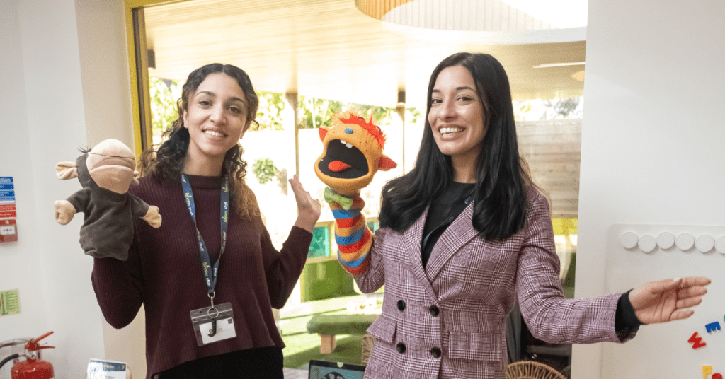 Two women in an office setting, one holding a puppet and the other a plush toy, smiling and posing playfully for the camera.