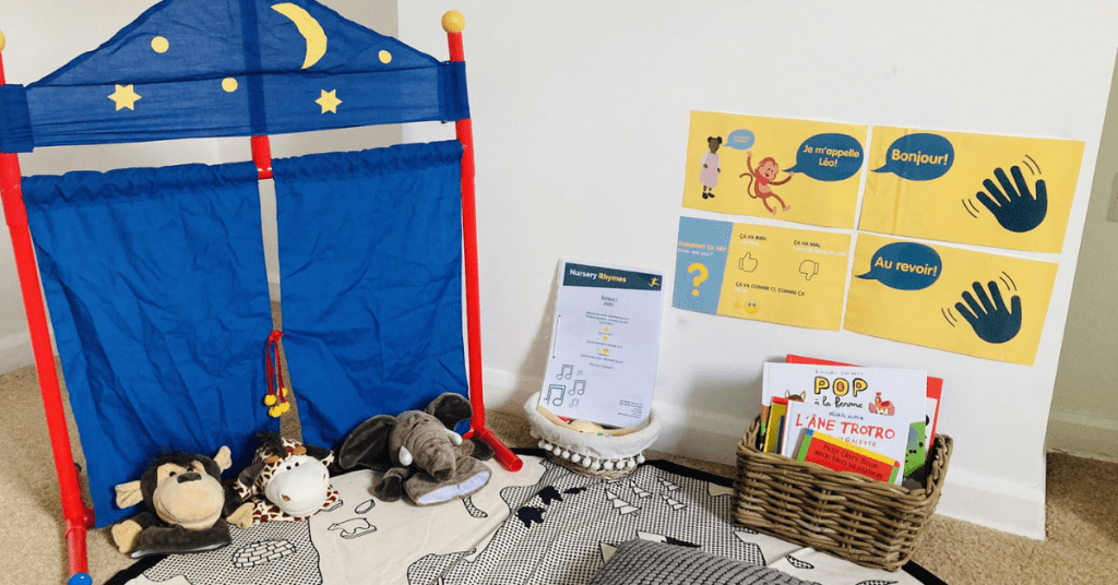A cozy child’s reading nook featuring a small blue puppet theater, a floor cushion with stuffed animals, books in a basket, and educational posters on the wall with language learning themes.