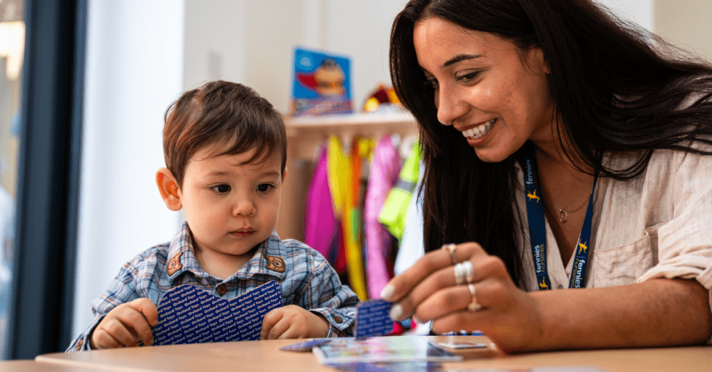 A young child and a smiling adult woman sit at a table, where the woman assists the child with a colorful learning activity. bright educational materials are visible in the background.