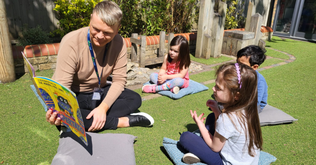 A teacher reading a book to three young children sitting on mats in a sunny garden, surrounded by wooden planters and greenery.