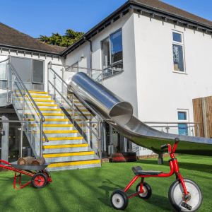 A modern white house with a large stainless steel slide attached to a balcony with yellow stairs. a red tricycle is parked on lush green artificial grass in the foreground.