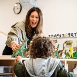 A joyful teacher engaged in play with a toddler at a classroom table full of colorful dinosaur toys. the teacher is animatedly opening her mouth wide, mirroring a dinosaur's roar, captivating the child's attention.