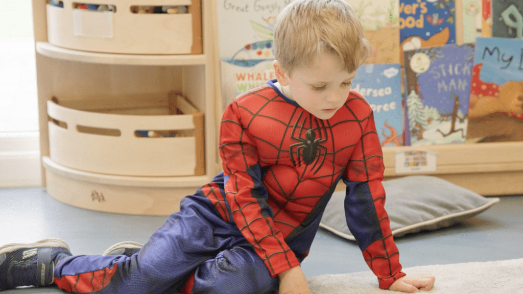 A young child dressed as spider-man kneels on a classroom floor, looking intently at something on the ground. the background features wooden shelves with books and toys.