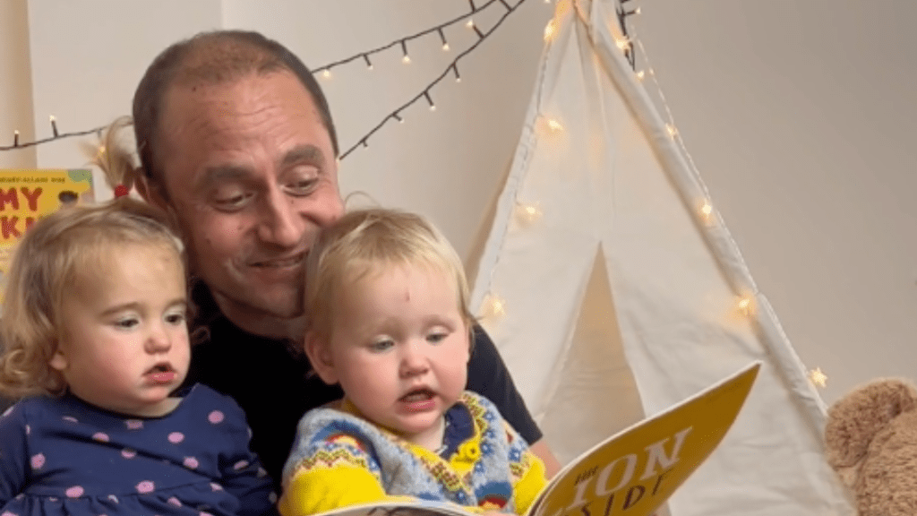 A man reading a book with two young children in a cozy room with a teepee tent and string lights in the background.