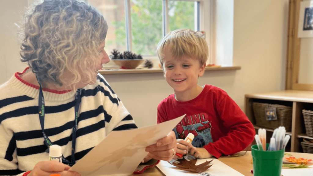 A young boy with blond hair smiling at a woman with curly hair as they look at a paper together in a classroom setting filled with art supplies.