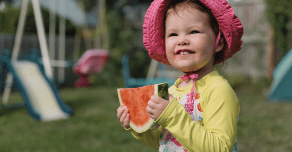 A toddler in a pink sunhat enjoys a slice of watermelon in a sunny backyard with a swing set in the background.