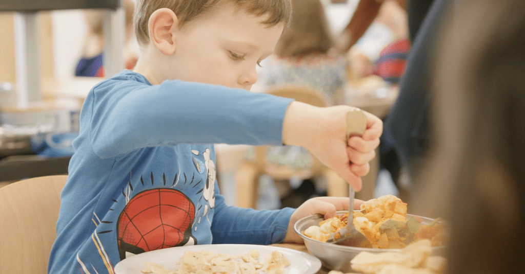 A young boy in a spider-man shirt eating from a bowl in a cafeteria, focusing intently as he uses his fork.