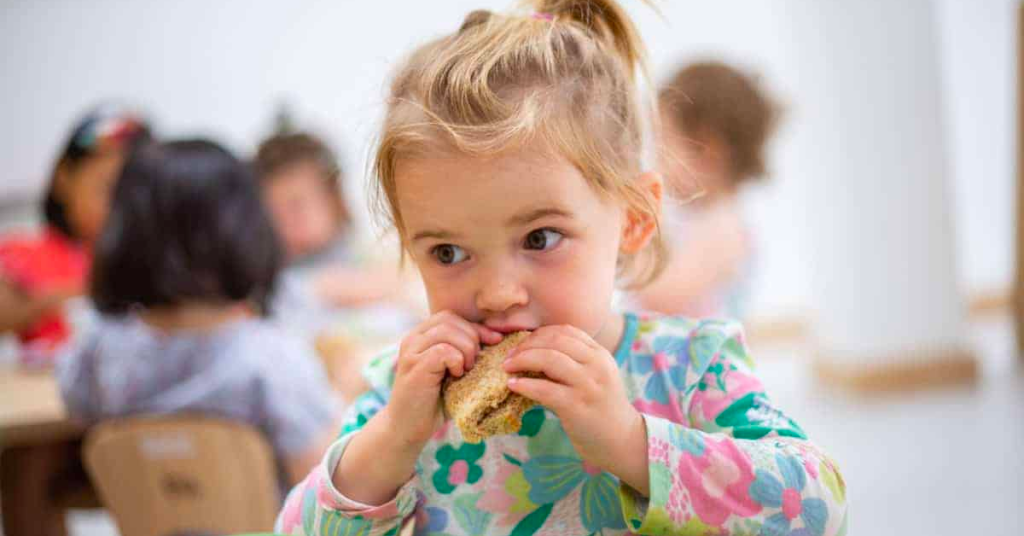 A young girl with blonde hair, wearing a floral shirt, eating a sandwich in a classroom with other children in the background.