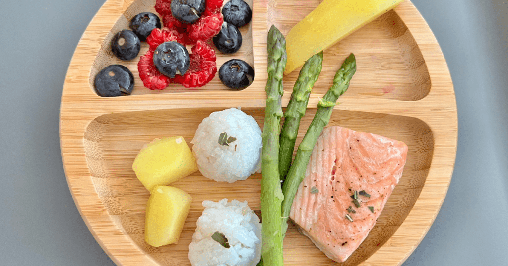 A wooden plate with a healthy meal consisting of grilled salmon, rice balls, asparagus, mango slices, and a mix of blueberries and raspberries.