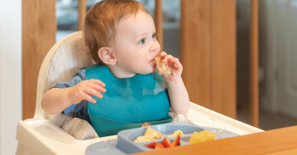 A toddler in a blue bib sits in a high chair, eating from a plate of fruit and bread.