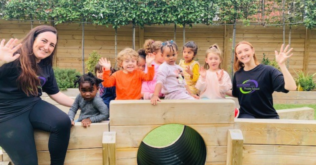 Two women and six children wave happily from a wooden play structure in a garden setting, conveying a cheerful and playful atmosphere.