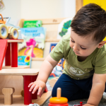A young child playing with toys in a colorful classroom, focusing intently on a wooden toy structure.