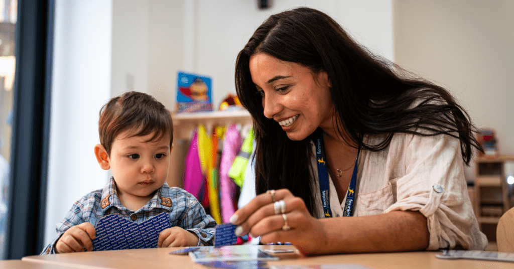 A woman and a young boy engage in learning activities at a table, with the woman smiling joyfully as she shows something to the attentive child. bright indoor setting with educational materials visible.