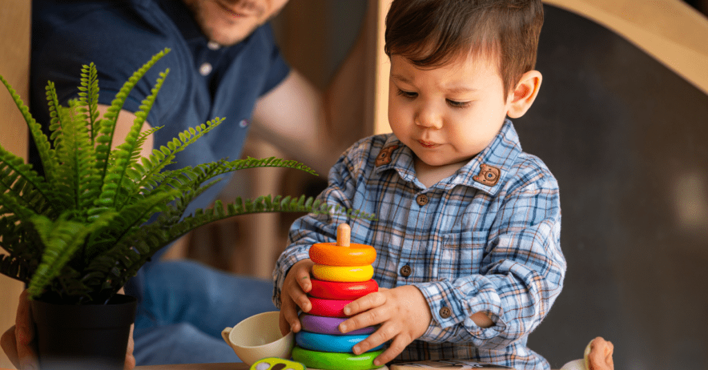 A toddler boy, focusing intently, stacks colorful rings on a spindle at a play table, with a male adult partially visible in the background.