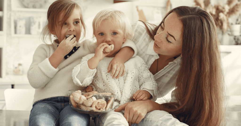 A mother and her two young daughters enjoy eating pastries in a cozy kitchen, sharing a moment of joy. the eldest daughter eats eagerly while the youngest is held by the mother.