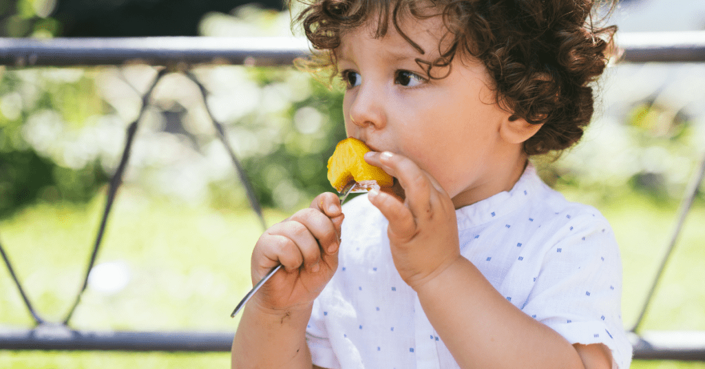 A young child with curly hair, wearing a white polka-dot shirt, intently eating an orange slice outdoors near a fence with greenery in the background.