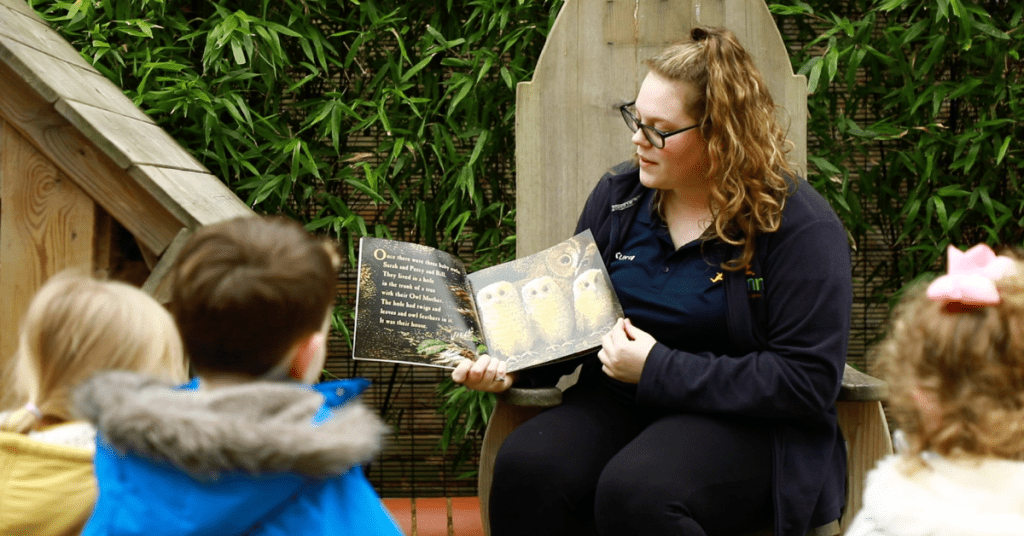 A woman with glasses, wearing a blue shirt, reads a book to young children in an outdoor setting surrounded by green plants.