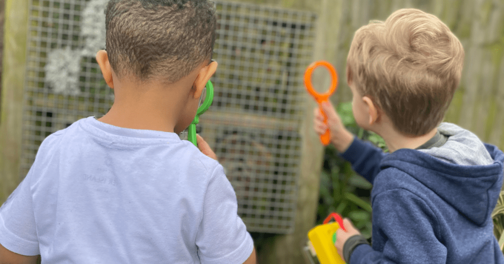 Two young children, one with brown skin and curly hair and the other with blonde hair, are viewed from behind. they are looking at a fence, holding magnifying glasses and a yellow bucket.