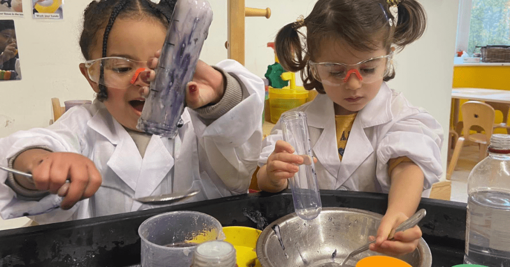 Two young girls wearing lab coats and safety goggles engage in a science experiment, mixing substances in a classroom setting. one examines a test tube closely while the other stirs a bowl.