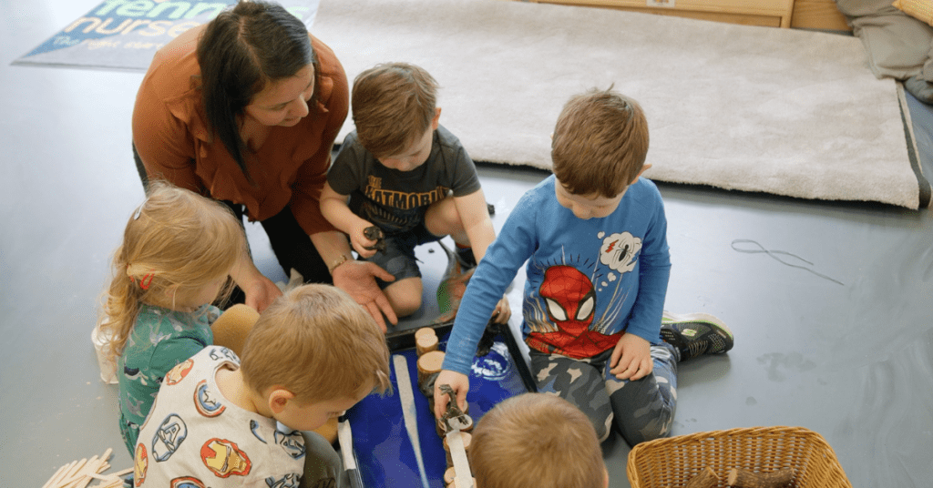 A teacher and young children engage in a playful activity on a classroom floor, surrounded by toys and learning materials.