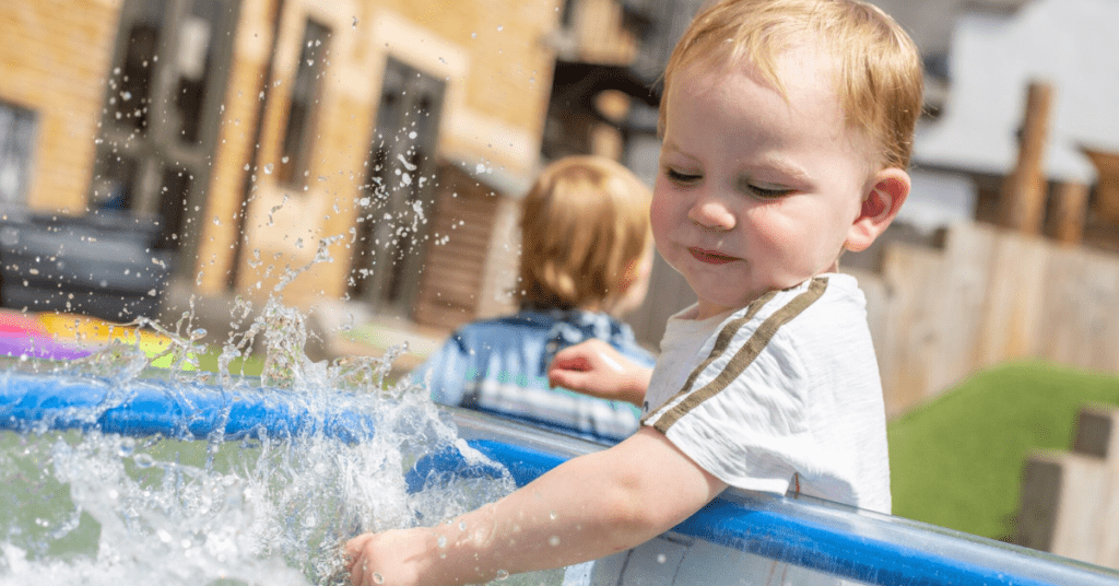 A toddler joyfully playing with water in an inflatable pool on a sunny day, splashing water around while another child is seen in the background.
