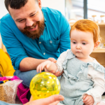 A toddler interacts with a colorful sensory ball, guided by a smiling man in a playroom, while another person sits opposite them, participating in the activity.