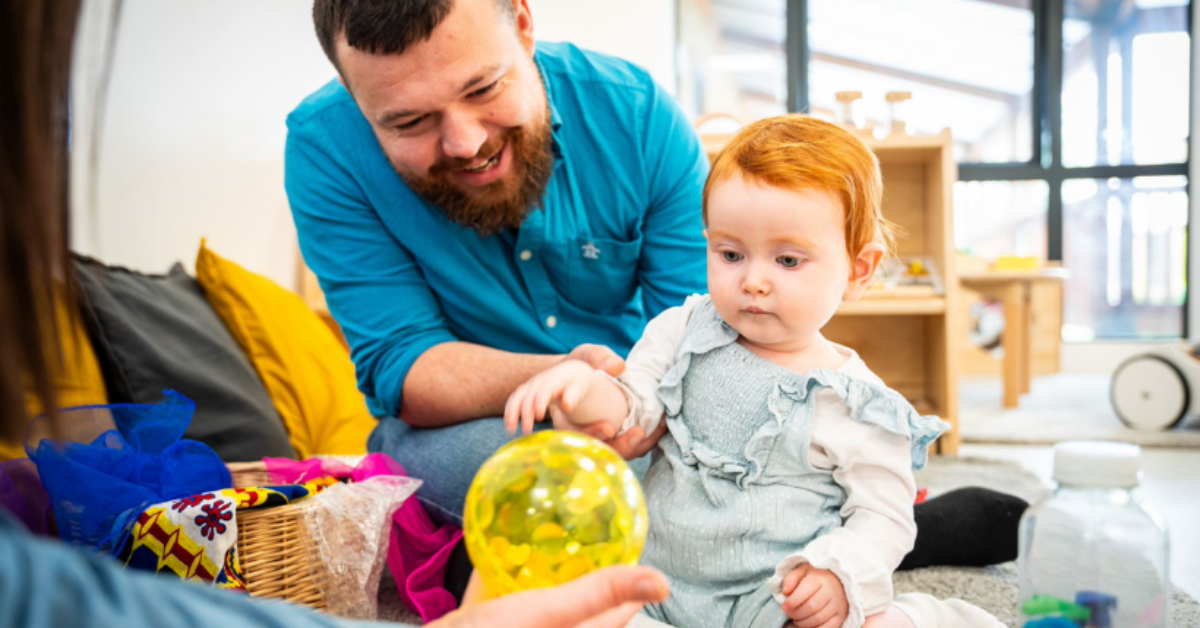A toddler with red hair interacts with a colorful sensory ball, guided by a smiling bearded man in a playroom, while another person sits opposite them, participating in the activity.