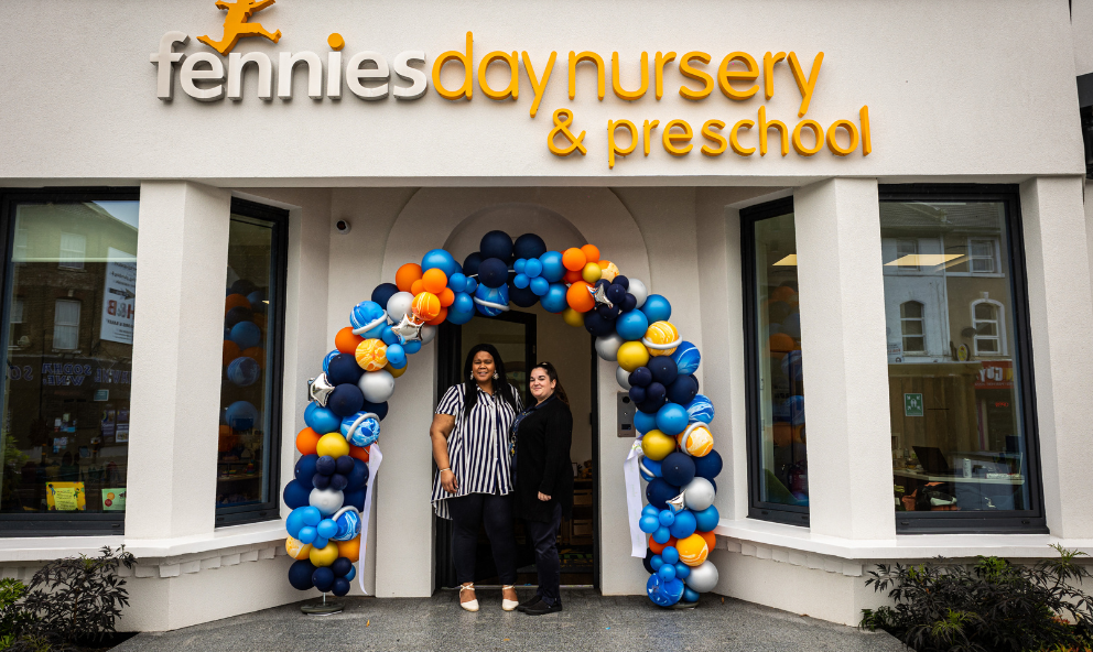 Two women smiling and standing under a blue and orange balloon arch at the entrance of 'fennies day nursery & preschool'. the building's façade is light-colored with a large window.