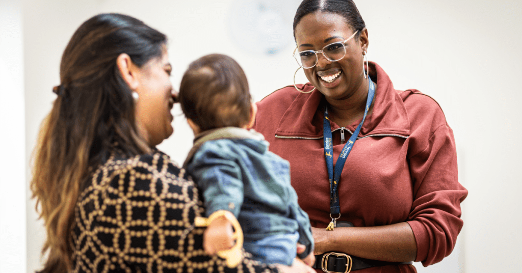 A professional woman wearing a lanyard smiles at a baby held by another woman in a patterned dress. the setting suggests a friendly workplace or daycare environment.
