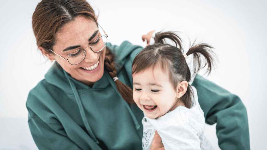 A smiling adult with glasses and a green hoodie holds a joyful toddler with pigtails. Both are looking at each other and laughing, creating a happy and playful moment. The background is a plain, light color, keeping the focus on their expressions.