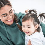 A smiling adult with glasses and a green hoodie holds a joyful toddler with pigtails. Both are looking at each other and laughing, creating a happy and playful moment. The background is a plain, light color, keeping the focus on their expressions.