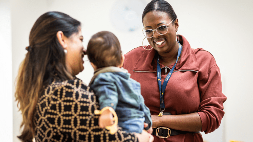 A woman holding a baby interacts with another smiling woman wearing glasses and a lanyard. The background is out of focus, emphasizing the warm interaction among the individuals.