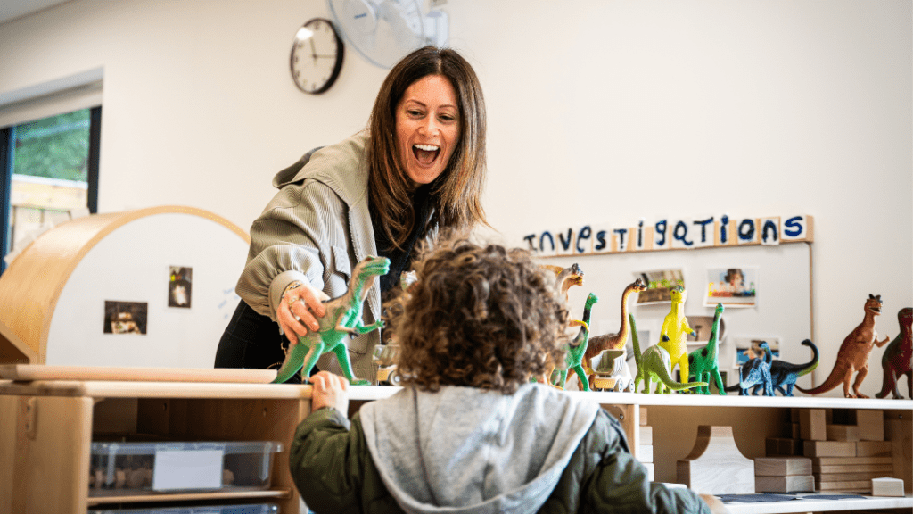 A woman and a child engage in playful learning with dinosaur toys in a classroom. The woman excitedly holds a green dinosaur, and the child watches intently. The background shows shelves with toys and decorations, including a sign that says "investigations.