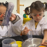 Two young girls wearing lab coats and safety goggles are engaged in a science experiment with beakers and colored liquids at a classroom activity table.