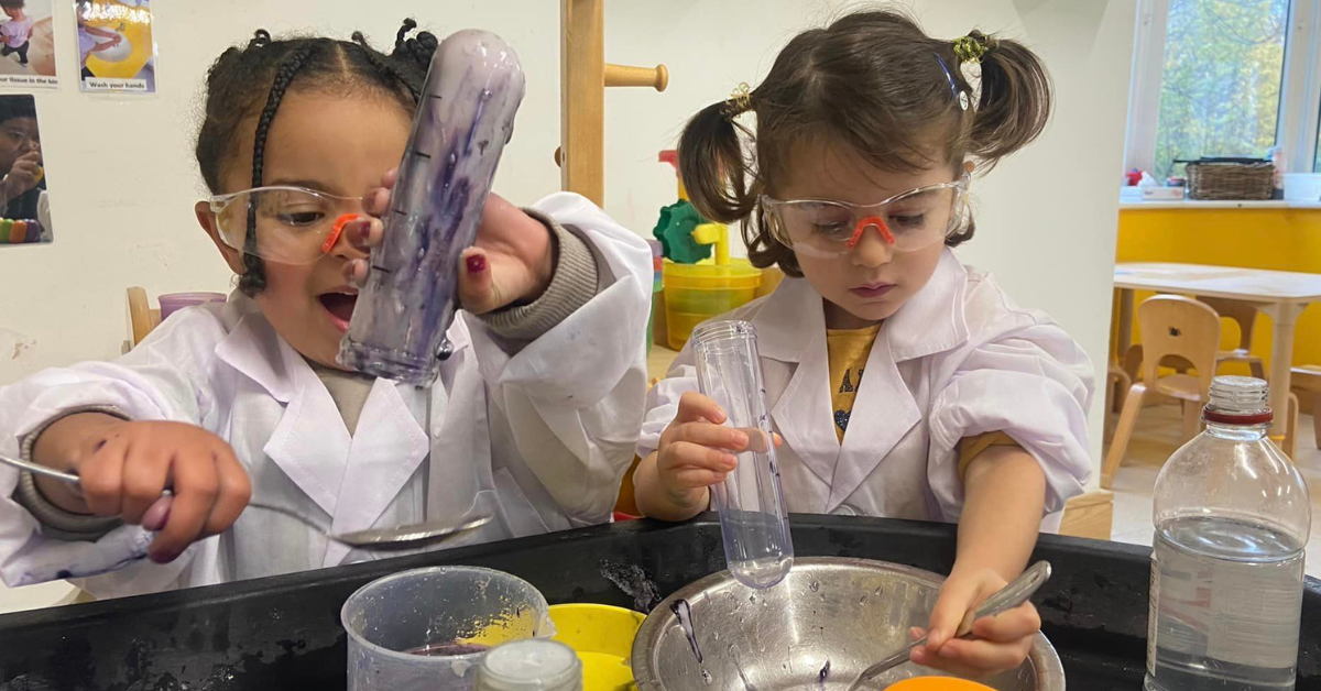 Two young girls wearing lab coats and safety goggles are engaged in a science experiment with beakers and colored liquids at a classroom activity table.