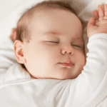 A baby with light skin is sleeping peacefully on its back, wearing a white outfit. The baby's right arm is raised near its head, and its facial expression is calm, with eyes closed and lips slightly parted. The background is soft and white.