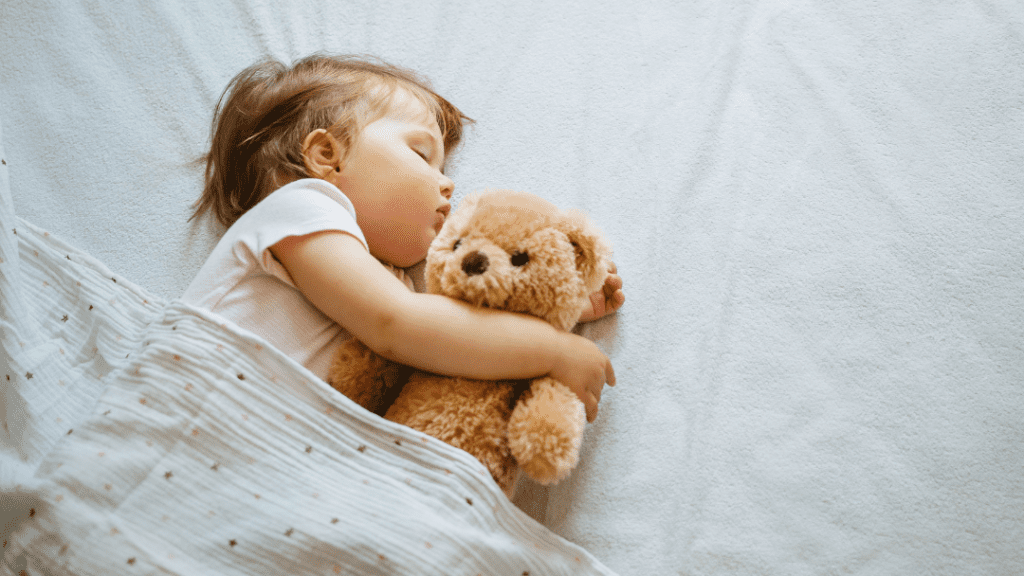 A young child sleeps peacefully on a white bed while hugging a brown teddy bear. The child is covered with a white blanket and wears a light-colored outfit. The scene is serene and comforting, reflecting a peaceful nap time.