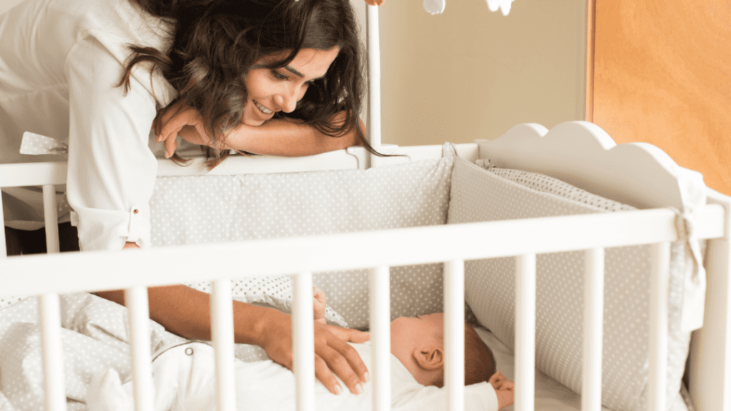 A woman with long, dark hair leans over a white crib, smiling at a baby lying inside. The baby is dressed in white, and the crib has soft, gray polka-dot padding. The scene is warmly lit, creating a cozy and loving atmosphere.