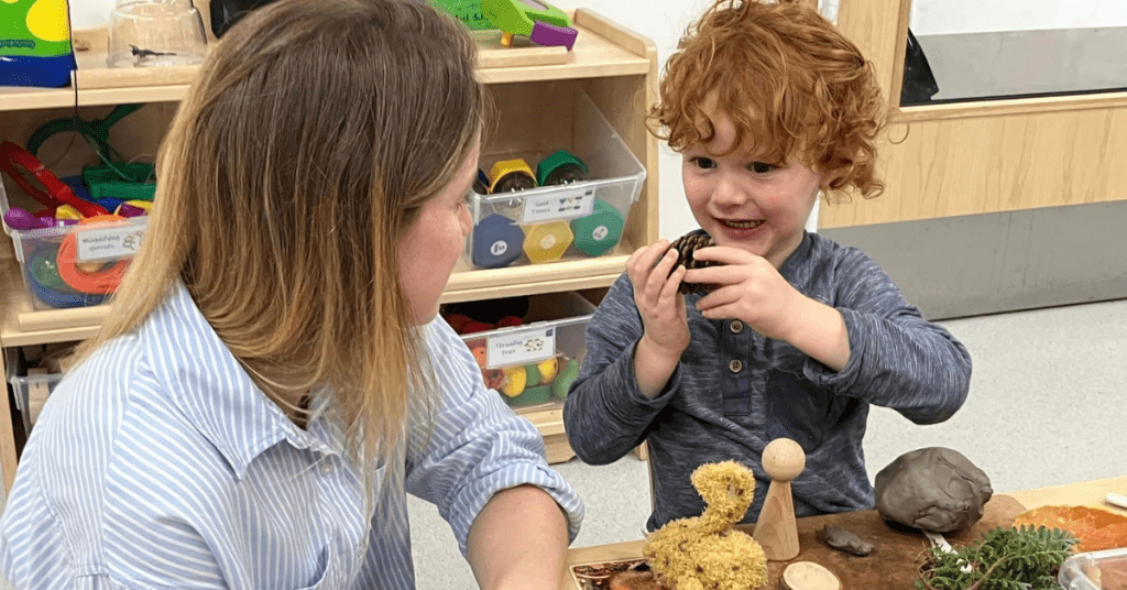 A young child with curly red hair holds an object and smiles, explaining it to a woman with blonde hair sitting across from him. They are at a table with various craft supplies, including a yellow pom-pom creature, a wooden figure, and modeling clay.