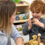 A young child with curly red hair holds an object and smiles, explaining it to a woman with blonde hair sitting across from him. They are at a table with various craft supplies, including a yellow pom-pom creature, a wooden figure, and modeling clay.