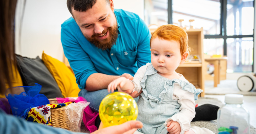 A bearded man in a blue shirt is sitting on the floor, smiling and looking at a red-haired child who is reaching for a yellow ball. They are indoors, surrounded by colorful toys and cushions. The atmosphere is playful and warm.