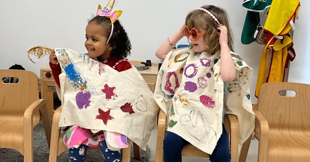 Two young children sit on wooden chairs, wearing handmade capes decorated with colorful shapes and stars. One child wears a unicorn headband, while the other wears heart-shaped sunglasses. They appear to be enjoying a playful activity together.