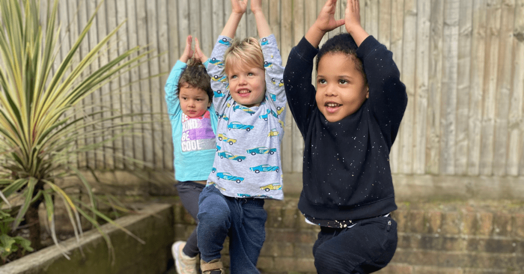 Three young children are standing on one leg with their arms raised above their heads in a yoga pose. They are outside, in front of a wooden fence and a plant. All three are smiling and appear to be enjoying the activity.