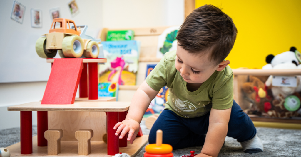 A young boy in a green shirt plays with a wooden toy truck on a playset in a brightly colored room. The room is decorated with various toys, books, and stuffed animals. The boy is focused on his play, with his hand reaching towards the toy set.