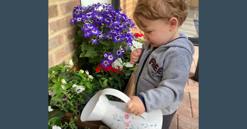 A young child with light brown hair, wearing a grey hoodie, stands on a brick patio and uses a white watering can with floral designs to water vibrant blue and red flowers in a wooden planter box against a brick wall.