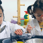 Two young children wear white lab coats and safety goggles while conducting a science experiment. They are focused on their task, holding various tools and containers filled with colorful liquids. The background features shelves with scientific equipment and bright classroom decor.