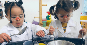 Two young children wear white lab coats and safety goggles while conducting a science experiment. They are focused on their task, holding various tools and containers filled with colorful liquids. The background features shelves with scientific equipment and bright classroom decor.
