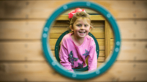 A young girl with a pink bow in her hair and a pink sweater with sparkly butterfly designs smiles while sitting in a wooden play structure. She is framed by a circular window with a teal trim. The background is blurred, focusing on her joyful expression.